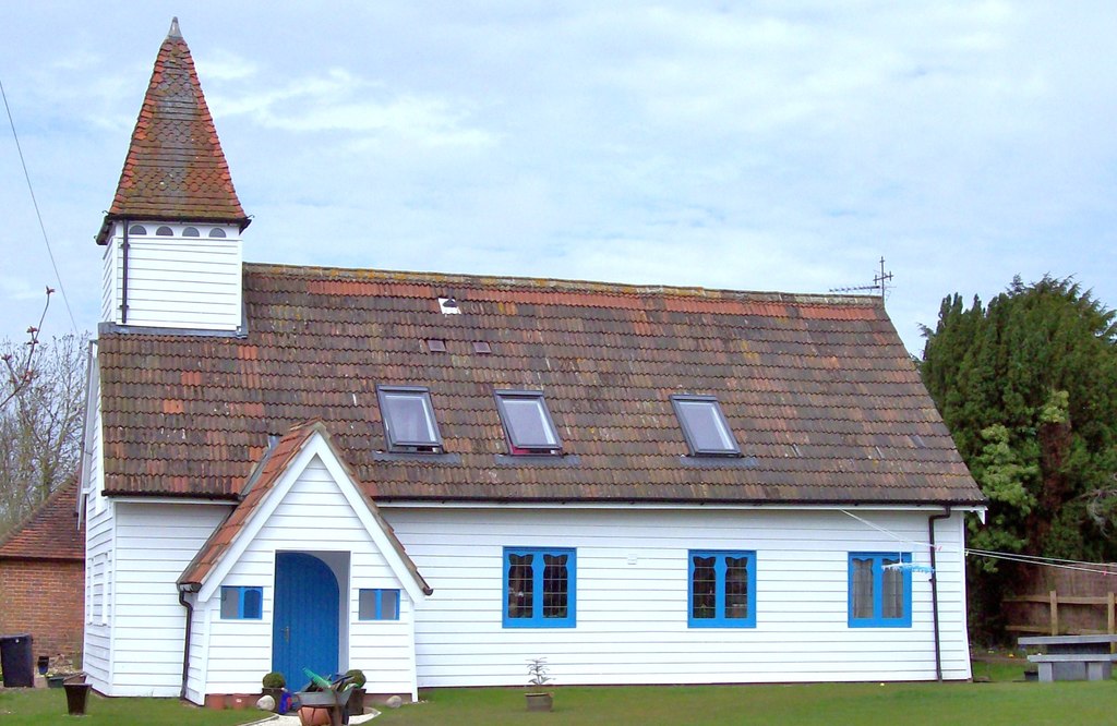 Quaintm small oblong church. White timber clad, with light blue window frames, and blue door. Small porch, and small bell tower above on left hand side on roof. Surrounded by vivid green lawn.