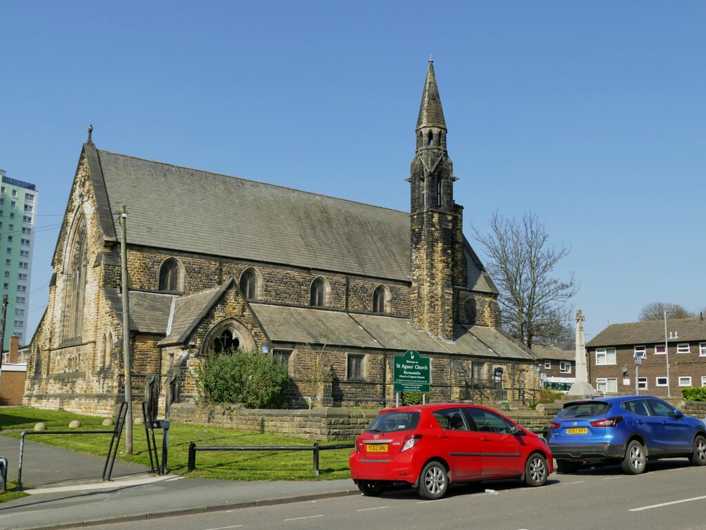 Church of Saint Stephen and Saint Agnes from the main road, on which are parked two cars in front of the church, one red and one blue. The backdrop is a gloriously clear blue sky, and one of the neighbourhood's tower blocks lurks in the background to the left of the shot. The church stretches across the photo from left to right, with a thin tower rising upwards on the right of the photo.