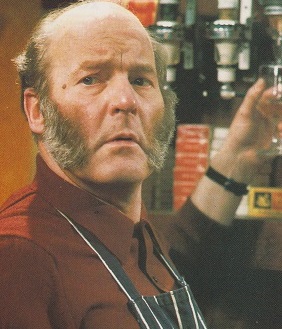 A colour cropped head and shoulders shot of the old Emmerdale Farm TV character Amos Brearly, reaching for the optics of a bottle behind the bar of the pub he worked in. He is showing off his trademark bushy sideburns and bald head, and wearing a dark apron with white stripes over a buttoned up burgundy coloured shirt.