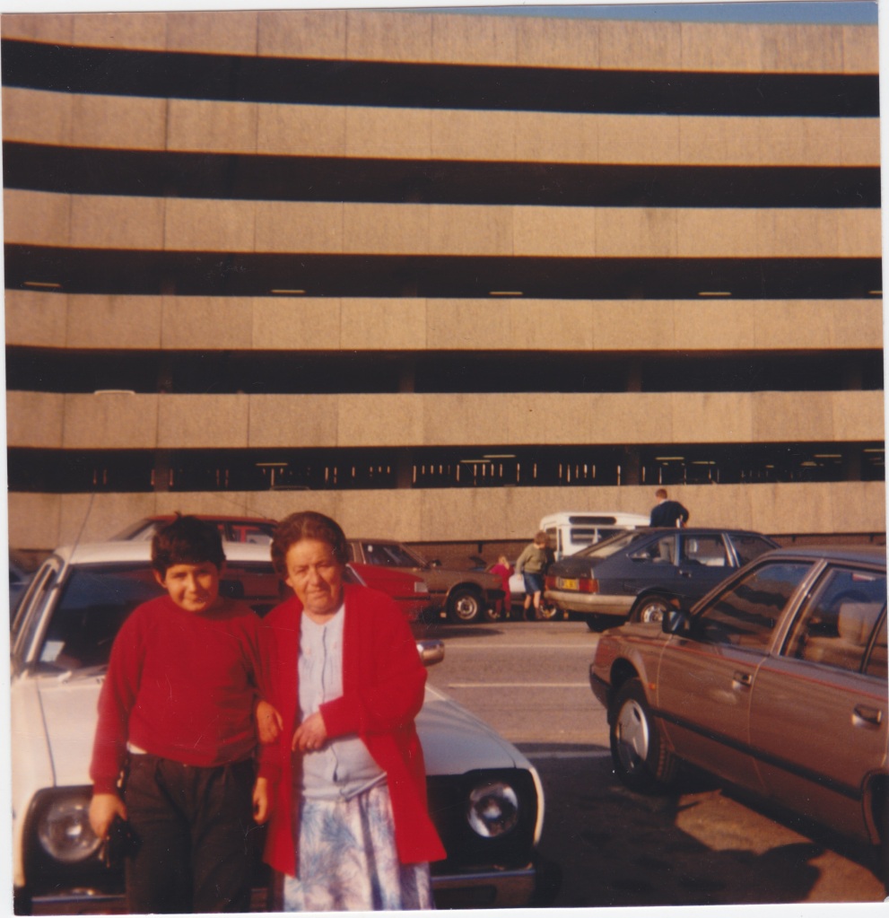 Colour photo of my young cousin arm in arm with our Nan, posing against the backdrop of a brutal multi-storey car park. They are sat against the bonnet of their white Datsun Sunny. A hint of bright blue sky across the top of the photo.
