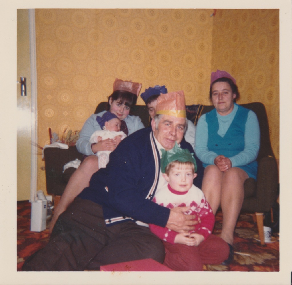 A bright colourful family photo at Christmas from the early 1970s. I'm sat on the floor with my Grandad in the foreground. Left to right on the sofa behind, my Mum holding my baby sister, my Dad obscured, and my Nan Joyce. Wallpaper of light orange dominates.