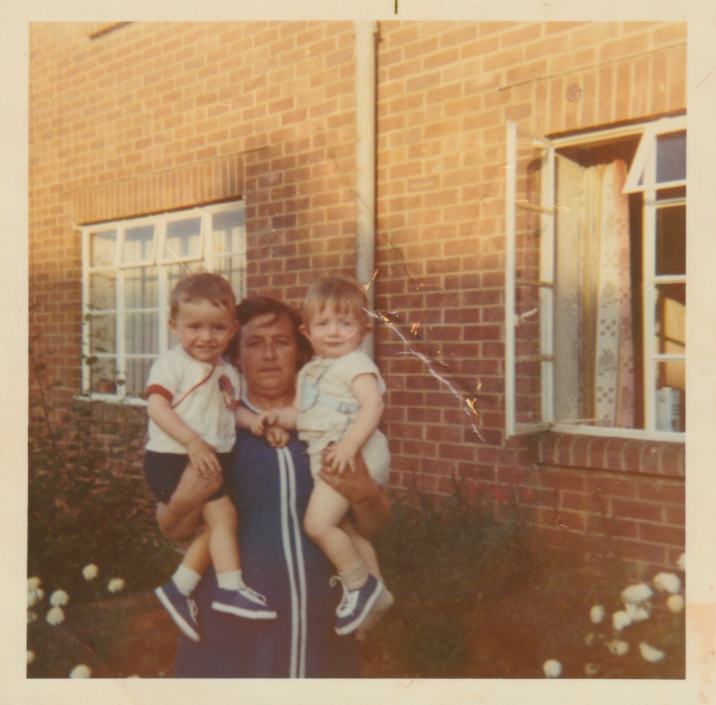Colour photo of the sun streaming onto the front of a council house in the seventies, my Nan in her early forties holding each of her first two grandsons in one arm for the camera. The borders in the garden are full of white flowers.