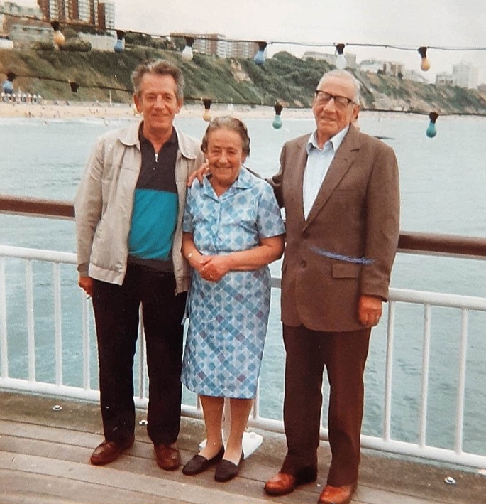 Colour photo of my Nan posing between two of her brothers on Bournemouth Pier, the coastline in the distance behind over the water and railing.