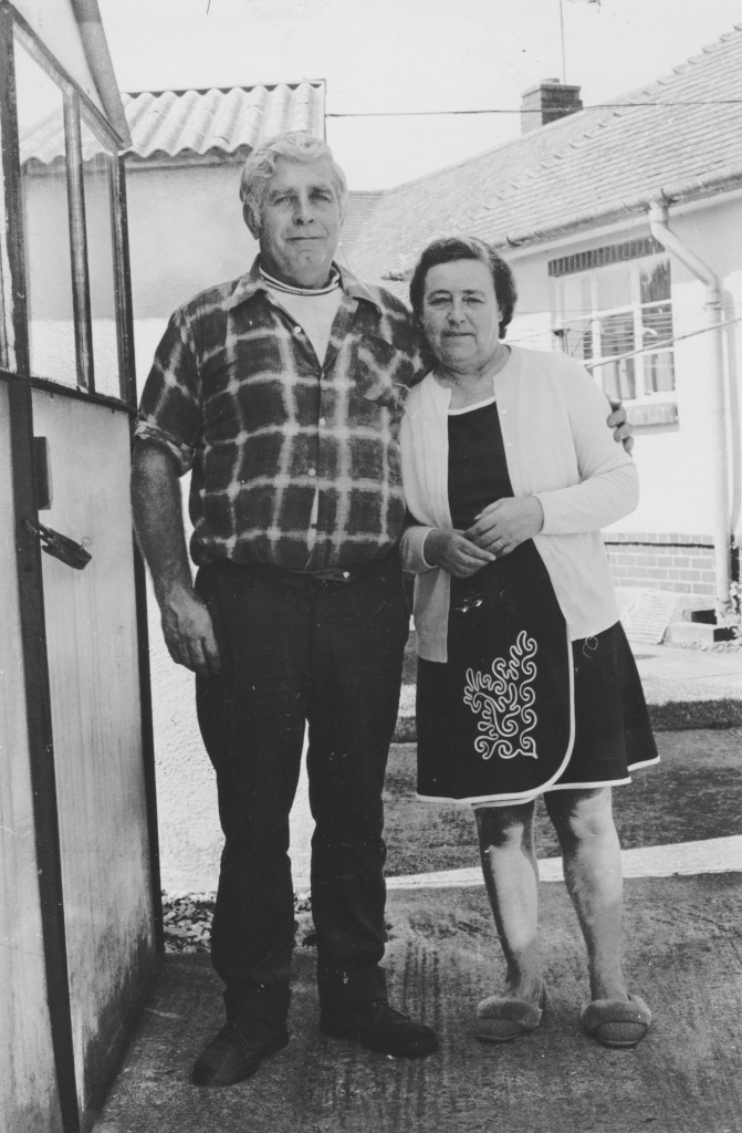 Black and white photo of my Mum's parents posing arm in arm, in front of their asbestos garage in the back garden to the left, with their neighbour's council bungalow in the background.