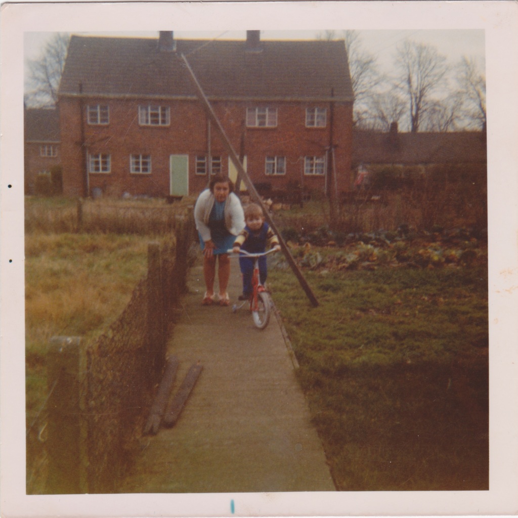 My Nan crouches behind me on the path in her slippers, giving me as a toddler all the support I need to learn to ride my red bicycle. To the left, behind a wire fence is the rough terrain of the neighbour's garden. To the right is the even rougher terrain of my grandparent's vegetable patch. In the background is the mirror image of their own council house, over the back fence.