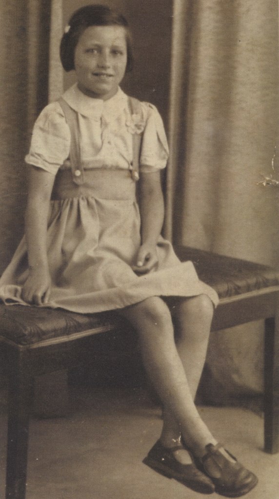 Sepia tinged photo of my Nan as a very young girl, sat on a very large stool, wearing sandals, legs dangling in the air. Formal looking photo, as if taken in a studio.
